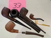 Six smoking pipes arranged on a surface including four regular sized wooden pipes with varying shapes and finishes plus two smaller miniature pipes.