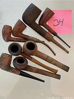 Seven assorted vintage smoking pipes laid out on white surface with a pink sticky note marked '34'
