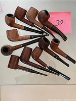Photo showing 11 used wooden smoking pipes with black stem mouthpieces, various bent and straight styles arranged on white surface with pink label showing number 30.
