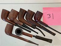Top-down view of seven wooden smoking pipes in varying shapes and sizes, arranged in row with pink paper marked '31'.