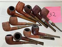 Photo of 11 wooden smoking pipes with various bowl shapes and lighter wooden stems, some with metal bands near bowls.