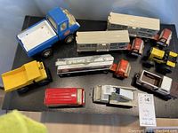 Overview of nine vintage Tonka trucks including horse vans, dump trucks, service van, oil tanker, and tractor on black platform