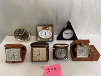 Overall view of all seven clocks and the clock repair book showing variety of shapes including triangular and rectangular, with brown and black leather cases.
