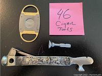 Three cigar tools including a cigar cutter, a vintage silverplate handled tool with farm scene relief, and a small chrome puncture tool on black surface.