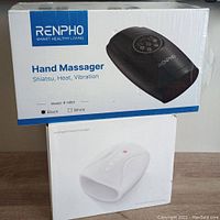 Two boxed hand massagers stacked, top is black Renpho Shiatsu hand massager box and below is white electric hand therapy massager box.