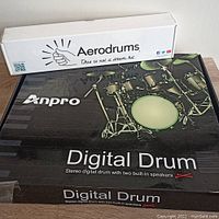 Boxes of Anpro digital drum kit and Aerodrums electronic drum set stacked together showing brand names and product type.