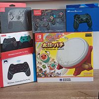Five boxed wireless controllers and one boxed Hori drum controller displayed on table