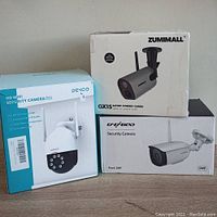 Photo showing all three security cameras in their original factory boxes: Zumimall GX1S, Dekco HD WiFi camera, and 1080P wired security camera.
