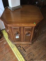 Front angle view of an octagonal light wood commode table showing the top surface, door with circular carved details, and base.