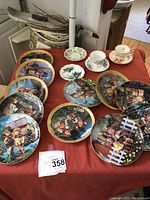 Top view of 11 Hummel plates showing colorful children scenes and 3 floral teacups with saucers on red cloth