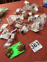 Wide view of multiple small bags containing various costume jewelry pieces arranged on a table. Pair of gold-toned shoe clips with orange rhinestones on leaf design in foreground on green card with price tag.