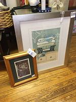 Photo showing two framed prints; the larger one is a stack of towels print with a silver-colored frame, the smaller one is a collectibles hutch print in a natural wood frame.