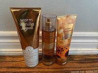 Front view of full size Bath and Body Works products: Warm Vanilla Sugar moisturizing body cream, Golden Sunflower body mist, and Golden Sunflower body wash arranged on floor in front of white baseboard and light wall.