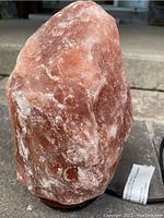Close-up image of the rough Himalayan salt lamp showing its natural pink-orange crystal with white mineral deposits and mounted on a base with part of the black electrical cord visible.