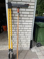 Three tools leaning against brick wall: post hole digger and squeegee tool at left, pump not shown