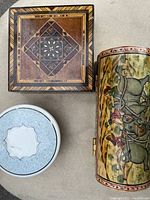 Top view of three decorative boxes: wood inlaid square box, painted cylindrical elephant box, and a small round white container