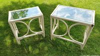 Two white rattan nesting tables with glass tops on grass outdoors showing size and curved design