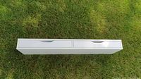 Top view of the white IKEA EKBY ALEX shelf with two drawers on a grassy surface, showing the drawer handles and overall shape.