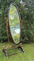 Oval mahogany swivel mirror with brass scroll side supports and wooden base stand, outdoor setting