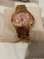 Close-up photo of rose gold ladies wristwatch with pink dial, chronograph subdials, date window, and linked bracelet on cushion.