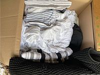 Box containing white queen bed sheets, black duvet cover, and various black and white towels with striped patterns.