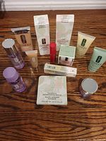 Overview of 13 Clinique products arranged on wood surface with boxes and tubes visible including bronzer powder, primer, foundation, and bottles.