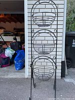 Front view of the decorative iron storage rack with three round wire baskets, placed outdoors against a light brick wall.