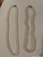 Two cultured pearl necklaces displayed side by side on a flat surface, showing their length, pearl size, and colors.