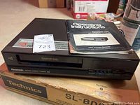 Panasonic VHS player showing front slot, brand and model labeling, with operating instructions placed on top, alongside original Technics turntable box beneath.