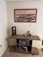 Photo of wooden table with clock, doll, terrarium object, CDs, and framed Indigenous art print above