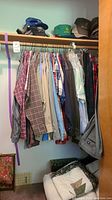 Men's casual shirts hung on a closet rod, a variety of colors and patterns, mostly plaid and stripes. Neutral tone hats are placed on the top closet shelf. Bottom shelf holds folded textiles including a quilt.