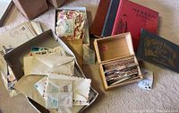 Photo showing wooden box filled with various postage stamps mounted on cardboard strips, envelopes with loose stamps, and two vintage stamp albums in red and blue covers on carpeted floor.