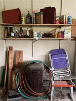 View showing aluminum extension ladder, shelves with oils and supplies, scrap wood planks, coiled air compressor hoses, metal container, and two purple folding chairs.