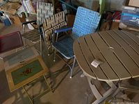 Photo showing seven foldable chairs with woven seats and backs in blue, brown, and red tones, plus two folding tables, one square with a green inset and one round white slatted patio table.