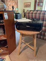 Canon Pixma MP495 printer on wicker stool next to wooden corner shelf unit with some papers on top.