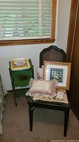 View of black wooden chair with woven backrest and floral cushion next to a small green side table with painted decoration, along with framed pictures and pillows placed on and around the furniture.