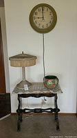 Photo showing the side table with lamp on top, wall clock hanging above, and planter on table.