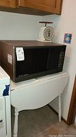 Black Sharp Carousel microwave oven placed on top of drop leaf table, with white kitchen scale on microwave.