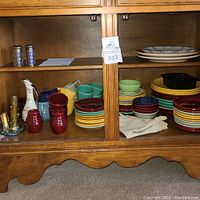 Wide shot of lower hutch shelf showing stacks of multicolor plates, bowls, mugs, doilies