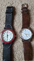 Photo showing front face of two wristwatches side by side on carpet, showing watch faces and bands.
