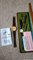 Two wristwatches side by side, one in a green velvet-lined box with accessories and paperwork, other without box.