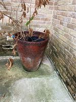 Large cone shaped ceramic pot with dried plant inside placed outdoors near brick wall, showing overall height and condition including visible crack.