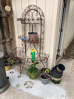Full view of the iron 3-shelf semi-circle plant stand showing ornate scroll design with pots hanging and placed on shelves and ground, with plastic containers and pots grouped near the stand.