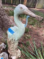 Close-up of a large heron statue with a weathered greenish finish, standing in an outdoor garden setting.