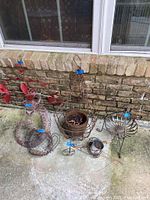 Six metal garden decorations arranged on patio floor against brick wall, includes lizard, bicycle planter, hanging basket, dragonflies, pitcher, and metal stand.
