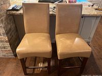 Two light brown upholstered barstools with wooden legs and footrests, shows wear and damage to upholstery and wood finish.