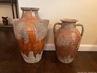 Two large clay pots with rustic patina and reddish-brown clay finish, displayed on hardwood floor against a white wall.