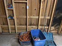 Wide view of lot showing extension cords, blue storage box, metal rake, and blue tool case against wooden garage wall