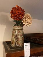 Tall distressed vase with two cream-colored and one burnt orange faux hydrangea blooms viewed from side