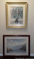 Two large framed watercolor prints hanging on a wall, one above the other. Top print is signed by Dolores Balsalabre with colorful abstract tree. Bottom print depicts a coastal sea scene.